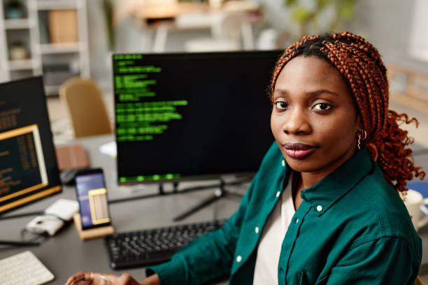 Portrait of young female IT developer looking at camera against programming code on computer screen in office interior
