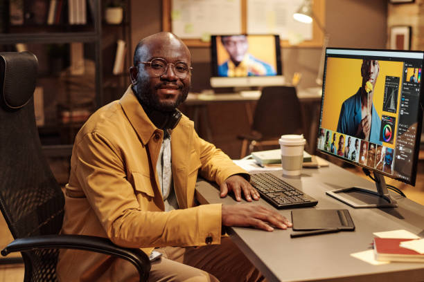 Portrait of African graphic designer in eyeglasses smiling at camera while working at table on computer with new content